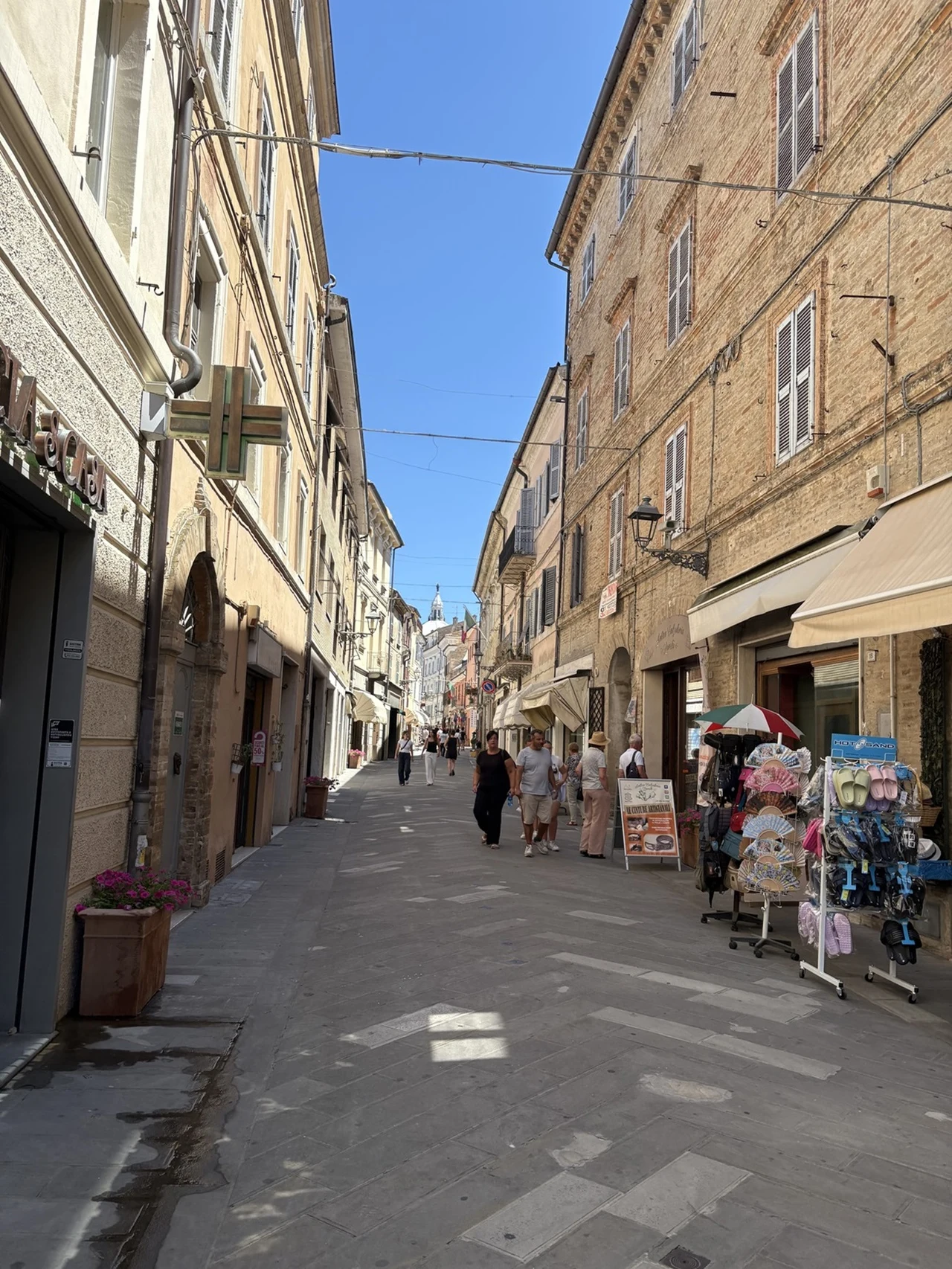 Wąska, brukowana ulica otoczona zabytkowymi kamienicami z jasnej cegły, po obu stronach znajdują się sklepy i stragany z towarami, a w tle widać przechodniów i fragment nieba.