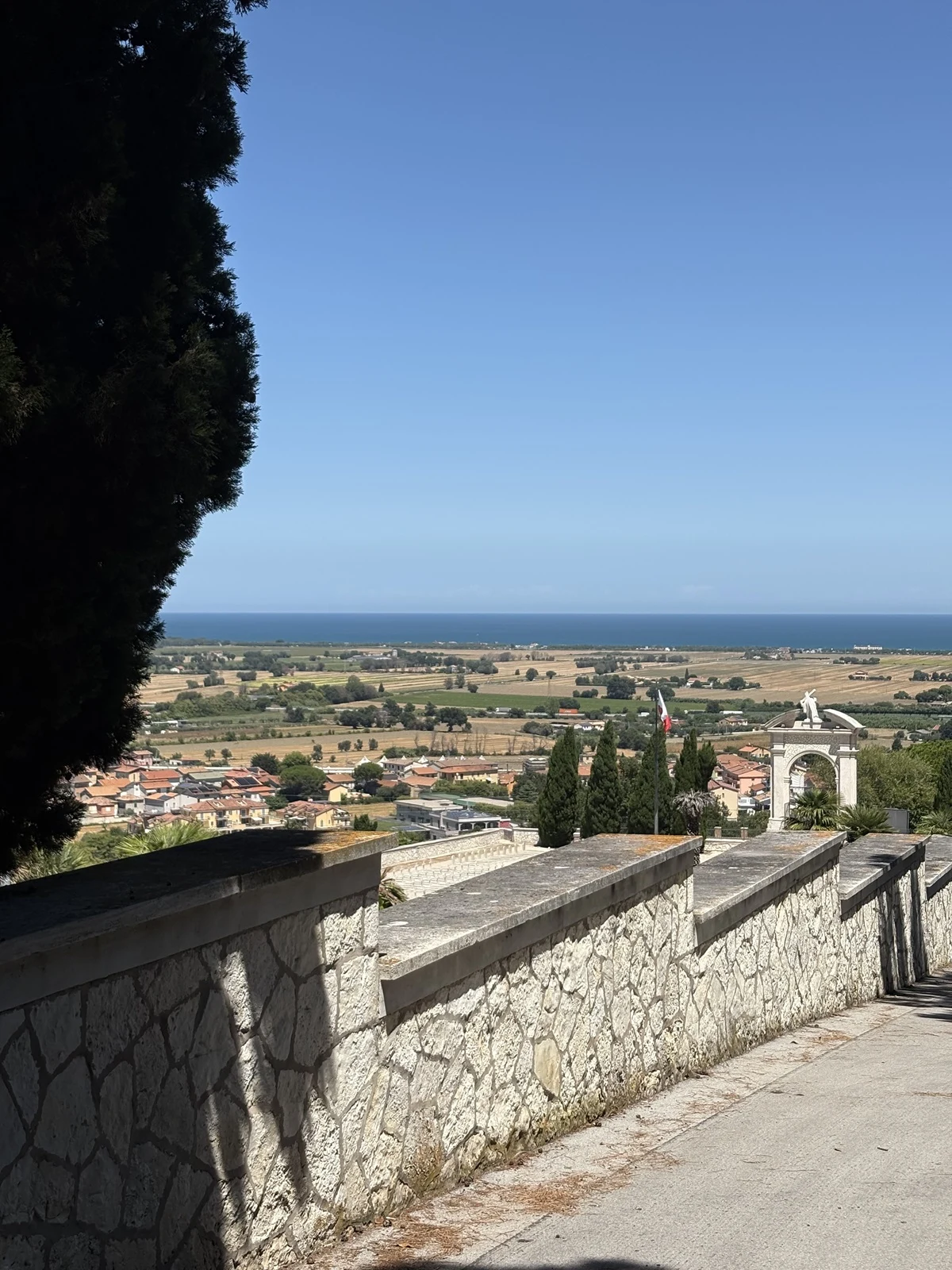 Kamienny mur prowadzący w dół do niewielkiego miasta, rozciągającego się w dolinie, z widokiem na szerokie pola i morze w tle. Nad krajobrazem dominuje niebieskie, bezchmurne niebo. Po lewej stronie widoczne duże drzewo, w tle charakterystyczny biały ł...