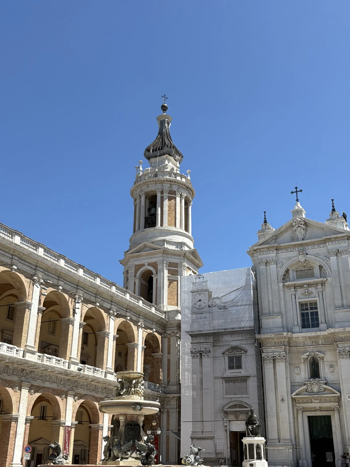Barokowa wieża bazyliki z kopułą i krzyżem położona obok klasycystycznego frontonu kościoła, ozdobiona licznymi detalami architektonicznymi. W lewym dolnym rogu widoczna fontanna z rzeźbami, a całość otacza arkadowy krużganek. Jasne, niebieskie niebo s...
