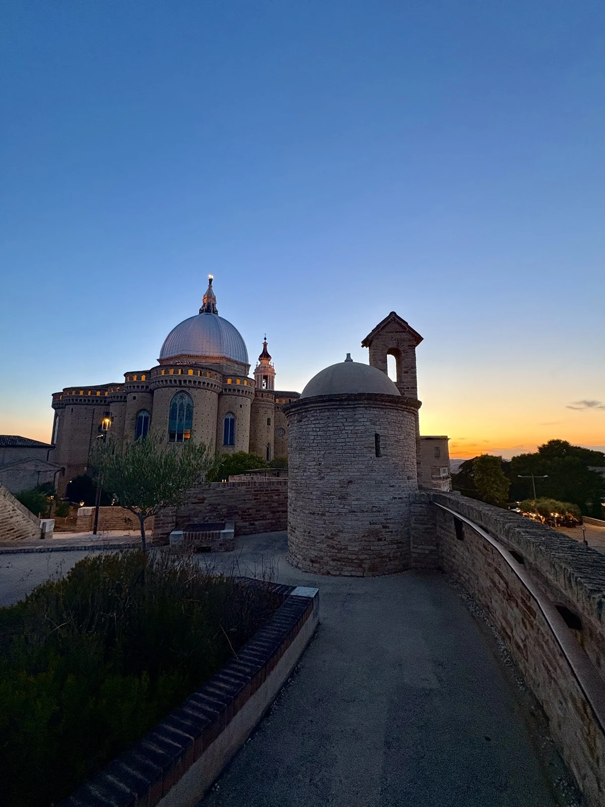 Kamienna kaplica z kopułą oraz dzwonnica otoczone historycznym murem, w tle większy kościół z dużą kopułą, całość oświetlona ciepłym światłem zachodzącego słońca, błękitne niebo bezchmurne.