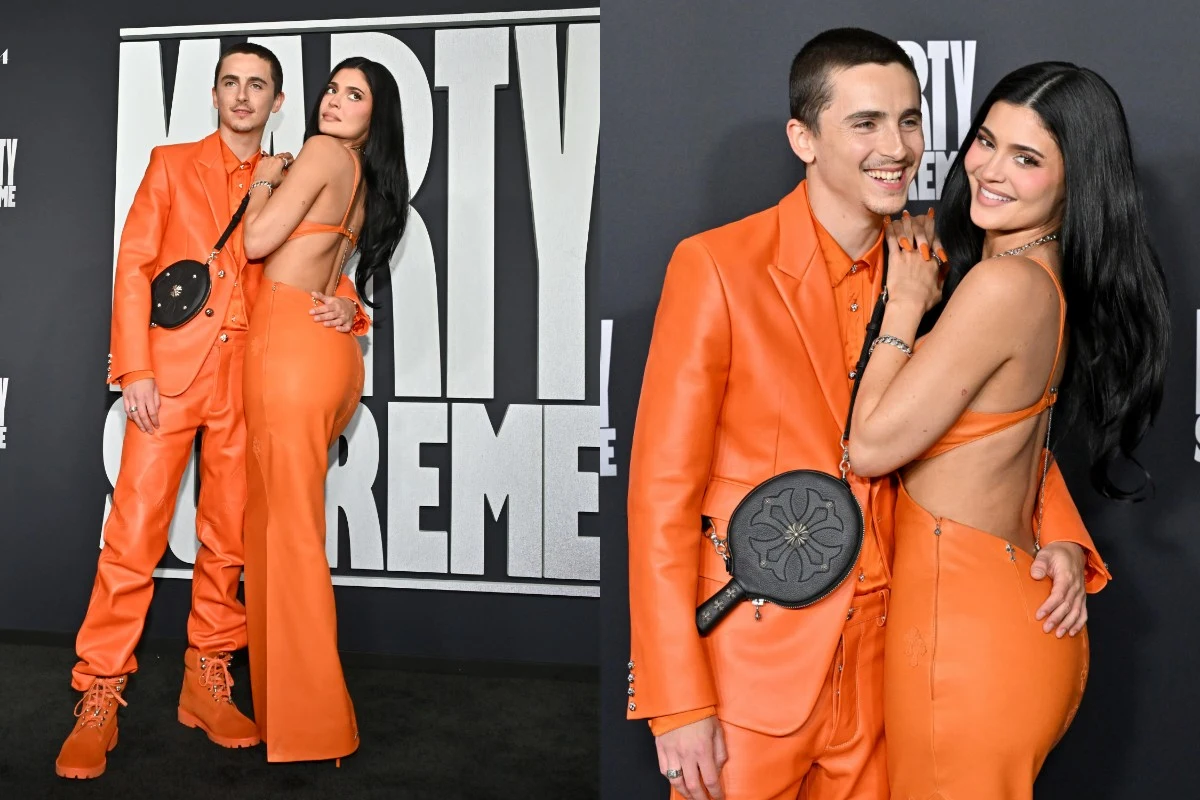 Para pozuje razem na tle ścianki eventowej, oboje ubrani w dopasowane pomarańczowe stroje. Kobieta ma długie, ciemne włosy i suknię z odkrytymi plecami, mężczyzna ma na sobie garnitur i sportowe buty. Obydwoje uśmiechają się oraz utrzymują bezpośredni ...