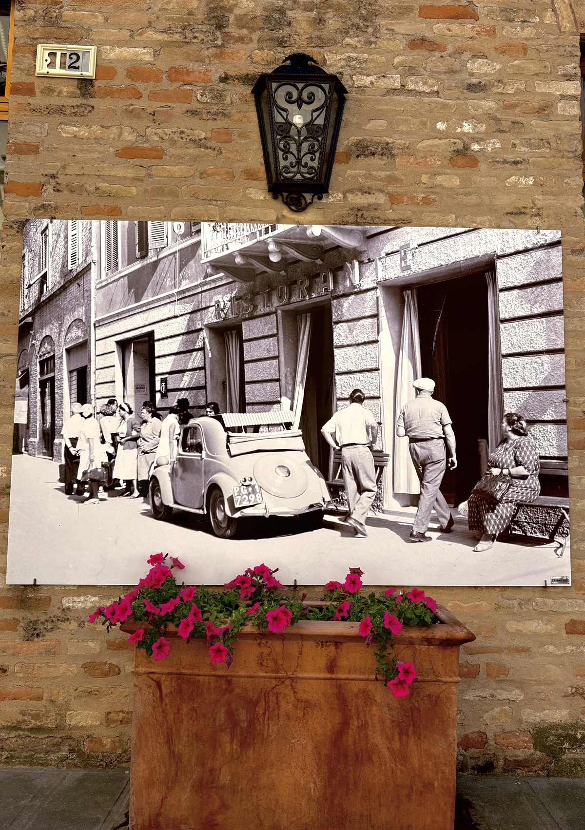 Stary czarno-biały plakat z widokiem ulicy, na której zaparkowany jest zabytkowy samochód, a wokół spacerują ludzie ubrani w stylu retro, umieszczony na tle ceglanej ściany z lampą oraz donicą z różowymi kwiatami.