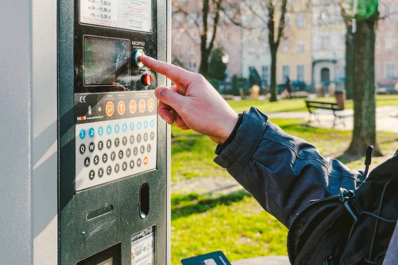 Darmowe parkowanie w płatnej strefie jest możliwe, pod warunkiem że znajdziemy przycisk "bułka" Osoba w kurtce naciska przycisk na parkometrze w parku miejskim, w tle ławki oraz drzewa i zabudowania mieszkalne.