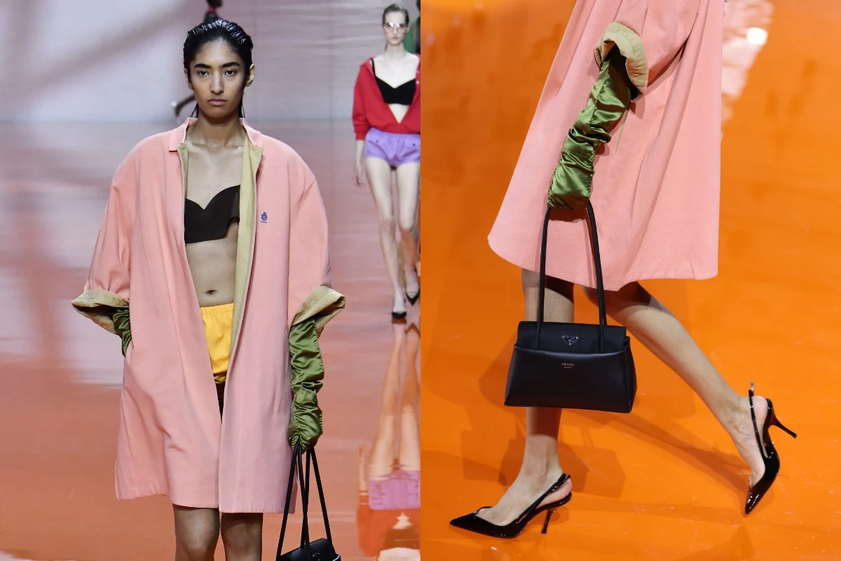 Młoda kobieta idąca po pomarańczowym wybiegu w oversizowym różowym płaszczu, zielonych rękawicach, czarnym staniku i żółtych spodenkach, niosąca czarną torebkę, w tle widoczna również inna modelka.
