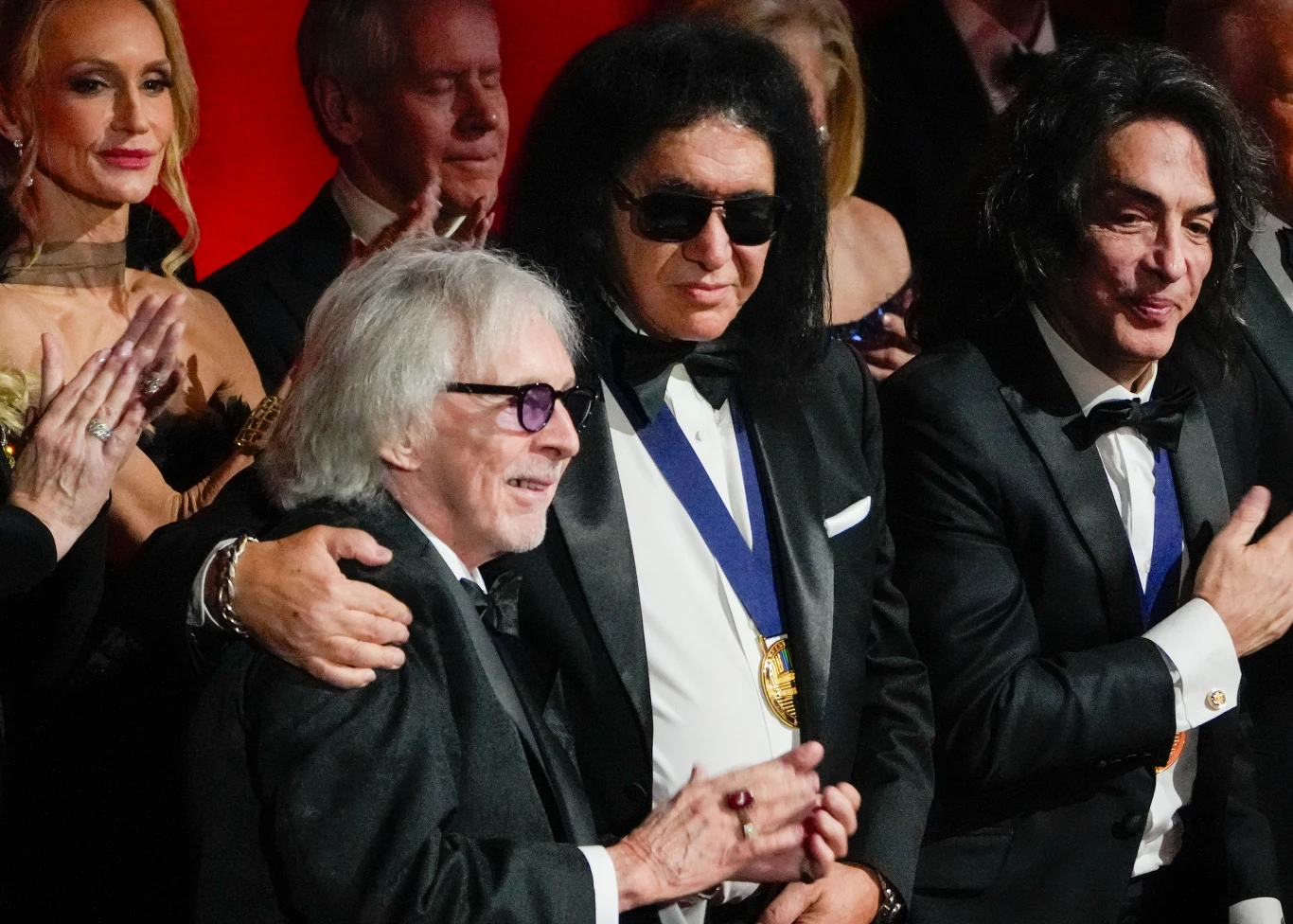 Peter Criss z liderami Kiss: Gene'em Simmonsem i Paulem Stanleyem podczas gali Kennedy Center Honors - 7 grudnia 2025 r. Kilku elegancko ubranych mężczyzn w garniturach i muchach stoi obok siebie na uroczystej gali, jeden z nich nosi ciemne okulary i medal na szyi, w tle widoczne są oklaskujące osoby.