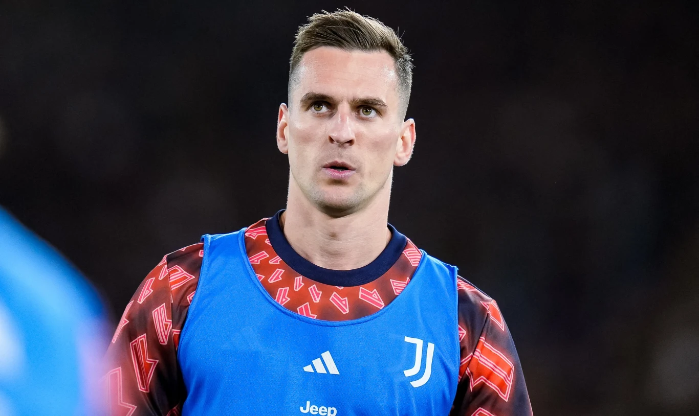 Piłkarz w niebieskiej kamizelce treningowej z logo Juventusu, skoncentrowany, w tle nieostre ciemne tło stadionu.