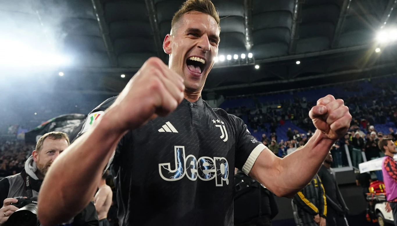 Piłkarz w koszulce Juventusu z uniesionymi pięściami wyraża ogromną radość na tle stadionu wypełnionego kibicami, wokół znajdują się osoby związane z drużyną i obsługą meczu.