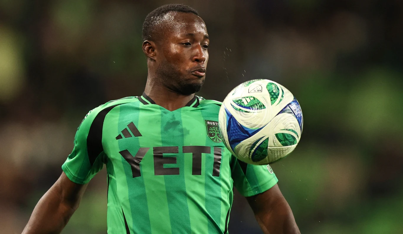 Piłkarz w zielonym stroju z logo YETI kontroluje piłkę podczas meczu, skupiony na grze, w tle rozmyte trybuny.