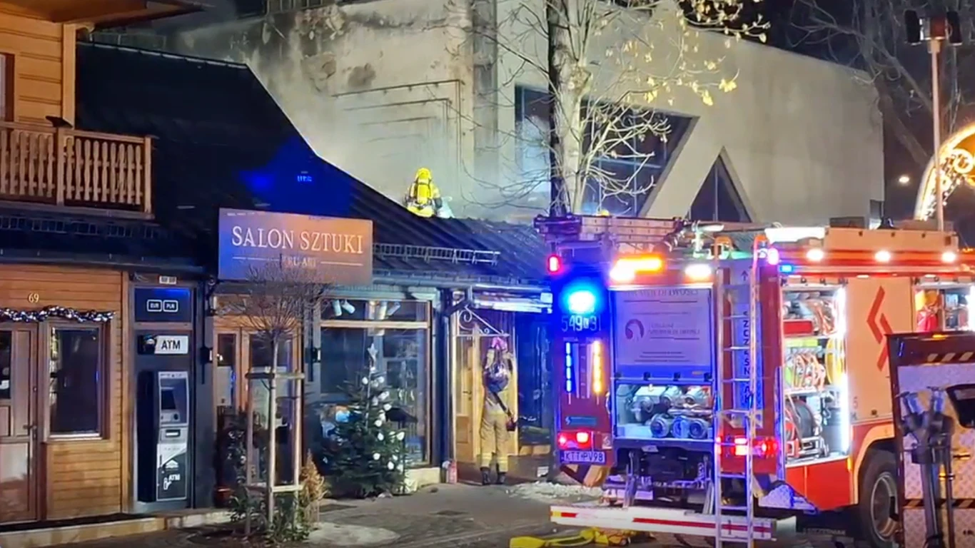 Zakopane. Ogromny pożar na Krupówkach. W ogniu stanęła drewniana restauracja Strażacy gaszący pożar budynku z szyldem 'Salon Sztuki', na pierwszym planie samochód strażacki z włączonymi światłami, na dachu widoczny strażak w kasku, wokół elementy otoczenia miejskiego.