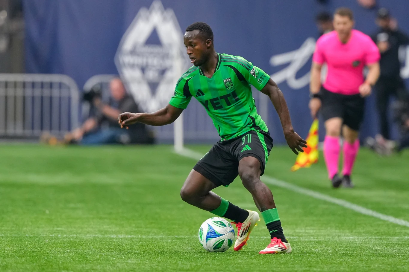 Piłkarz w zielono-czarnym stroju kontroluje piłkę na boisku podczas meczu, w tle sędzia w różowej koszulce oraz zarys kibiców i elementów stadionu.