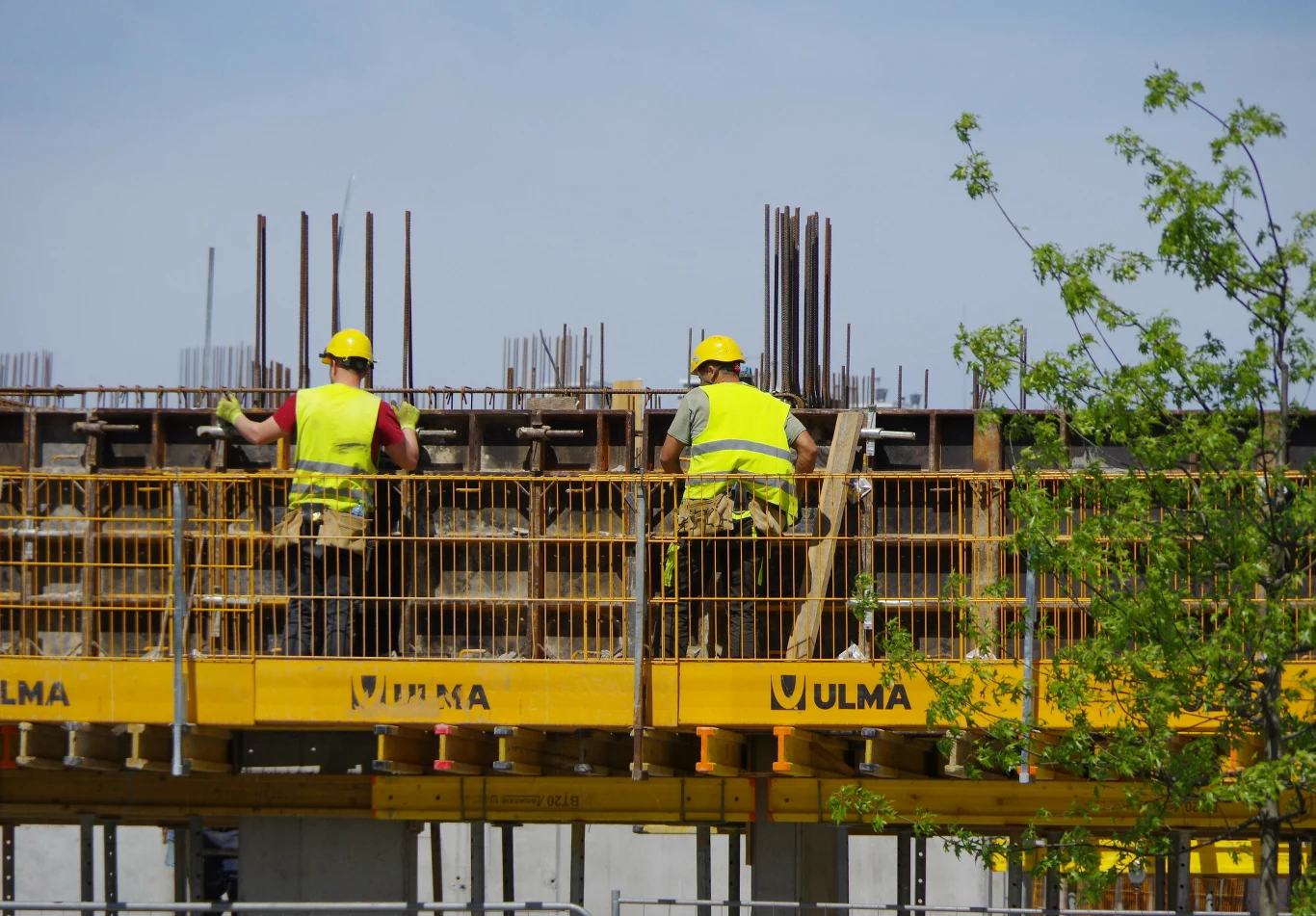 Nowe przepisy wchodzą od stycznia, dla wielu oznaczają 26 dni urlopu (zdj. ilustracyjne) Dwóch pracowników budowlanych w żółtych kaskach i kamizelkach odblaskowych wykonuje prace na wysokości przy szalunku nowo powstającego budynku, otoczeni metalowymi prętami zbrojeniowymi i żółtymi panelami konstrukcyjnymi marki Ulma.