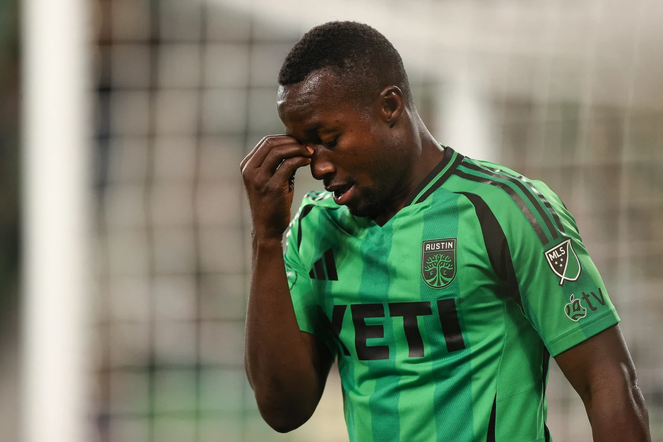 Piłkarz w zielonej koszulce z logo klubu Austin FC oraz sponsorów przeciera oczy w geście zmęczenia lub rozczarowania na tle bramki piłkarskiej.