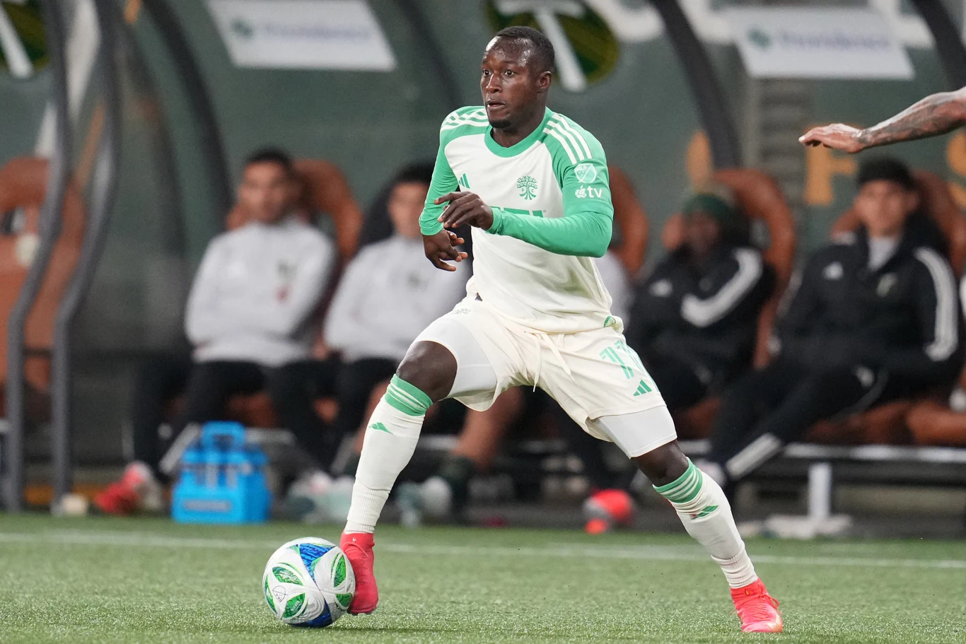 Czarnoskóry piłkarz w biało-zielonym stroju prowadzi piłkę na boisku podczas meczu, w tle widać ławkę rezerwowych i innych członków drużyny.