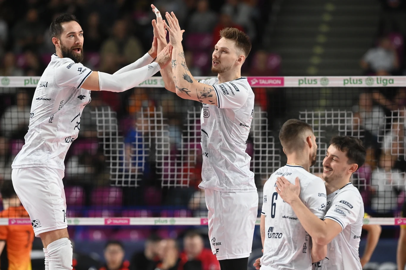 Grupa siatkarzy ubranych w białe stroje świętuje zdobycie punktu na boisku, wykonując wzajemne gesty radości. Atmosfera jest dynamiczna, a w tle widoczne są elementy hali sportowej oraz siatka.