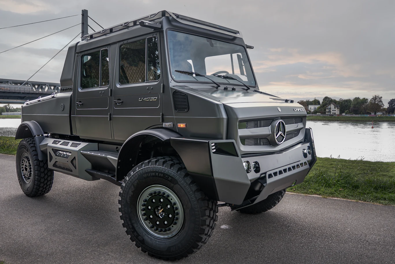 Samochód terenowy Mercedes Unimog U 4000 z charakterystycznym masywnym nadwoziem, terenowymi oponami i stalowymi osłonami, zaparkowany na asfalcie nad rzeką, w tle most i zieleń.