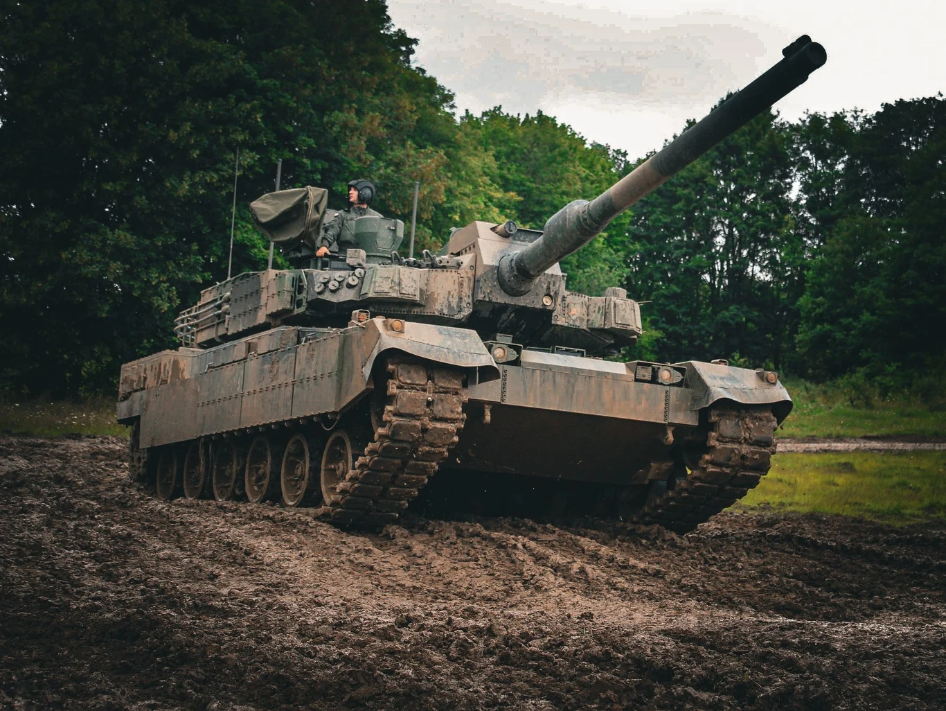 Czołg wojskowy poruszający się po błotnistej drodze na tle zielonych drzew, z widocznym żołnierzem w wieżyczce i podniesioną lufą.