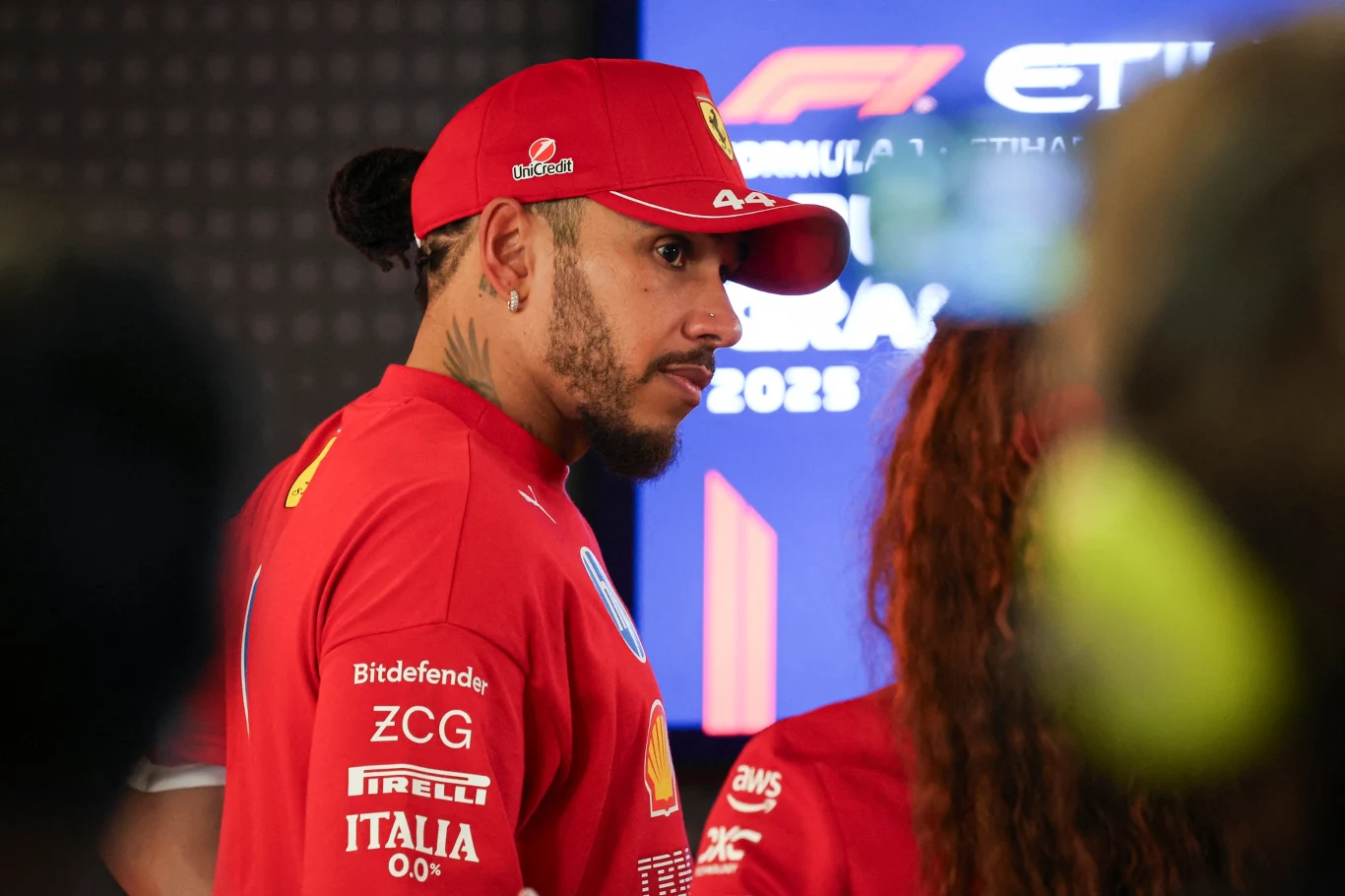 Kierowca zespołu Ferrari ubrany w czerwony strój sportowy i czapkę z daszkiem z logo zespołu, stoi na tle ekranu z grafiką nawiązującą do wydarzenia Formuły 1. Po prawej widoczna osoba w podobnym stroju, a wokół panuje atmosfera konferencji prasowej lu...