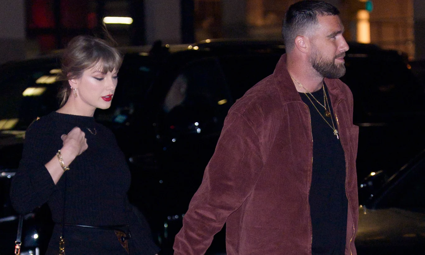 Taylor Swift i Travis Kelce Kobieta w czarnym stroju i mężczyzna w brązowej kurtce idą razem nocą po mieście, trzymając się za ręce. Za nimi ciemne samochody i rozświetlone budynki.