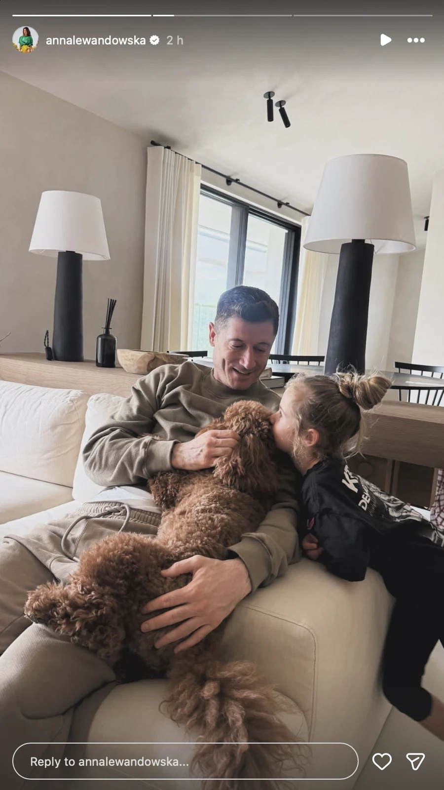 Mężczyzna siedzi na jasnej sofie w nowocześnie urządzonym salonie, obok niego dziecko przytulające się do brunatnego psa. W tle duże okna i dwie duże lampy stojące na komodzie pod ścianą.