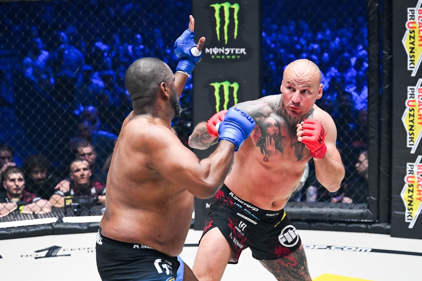 Dwóch zawodników walczących w oktagonie podczas gali MMA, jeden z nich wykonuje cios, drugi broni się podnosząc gardę, wokół tłum kibiców i charakterystyczne logo sponsorów na bandach.