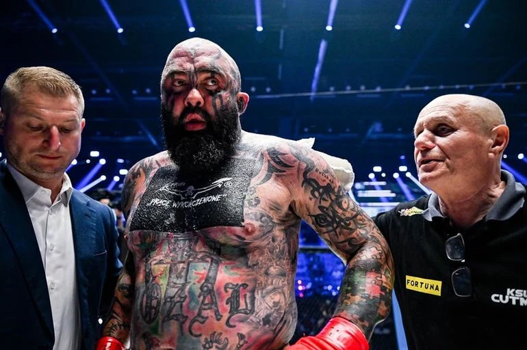 Zawodnik MMA z licznymi tatuażami na ciele, stojący na ringu po walce w otoczeniu dwóch mężczyzn, jeden ubrany formalnie, drugi w stroju sędziego sportowego, światła areny w tle dodają dramatyzmu scenie.