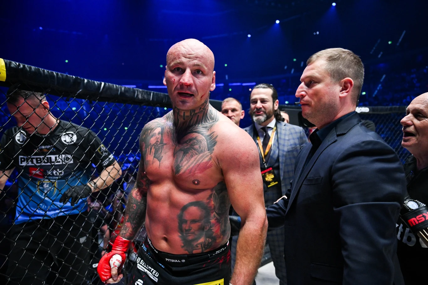 Zawodnik MMA z licznymi tatuażami na ciele stoi w klatce po zakończonej walce, wokół niego ekipa techniczna oraz ochrona, w tle rozświetlona arena z publicznością.
