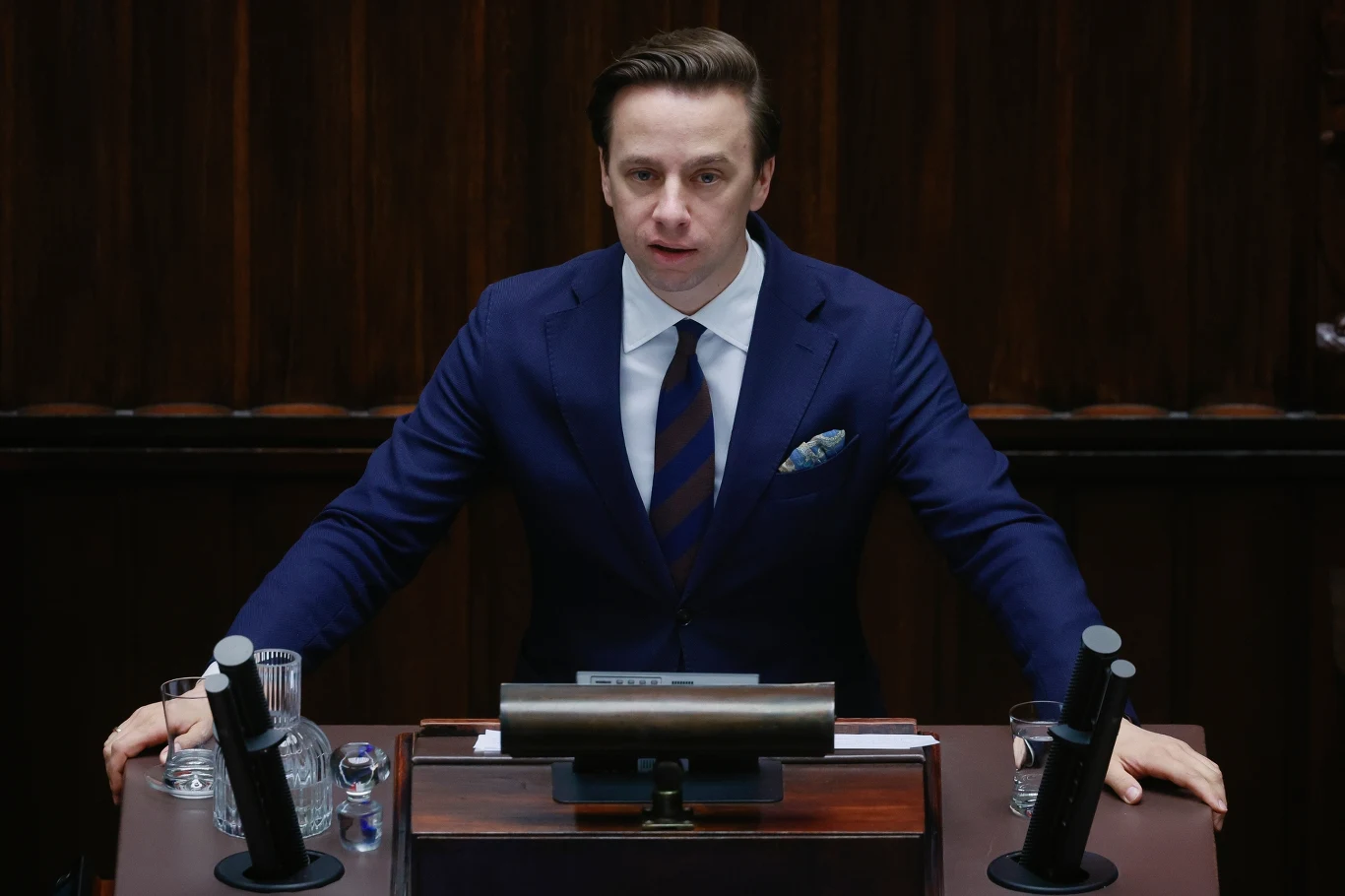 Mężczyzna w garniturze przemawia przy mównicy w sali parlamentarnej, na blacie znajduje się mikrofon i szklanki z wodą.