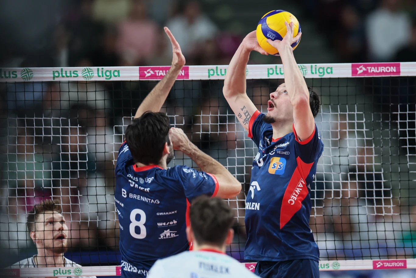 Dwóch siatkarzy drużyny w niebieskich strojach wykonuje akcję przy siatce, jeden podnosi piłkę do rozegrania, drugi przygotowuje się do ataku. W tle siatka, reklamy ligi oraz kibice na trybunach.