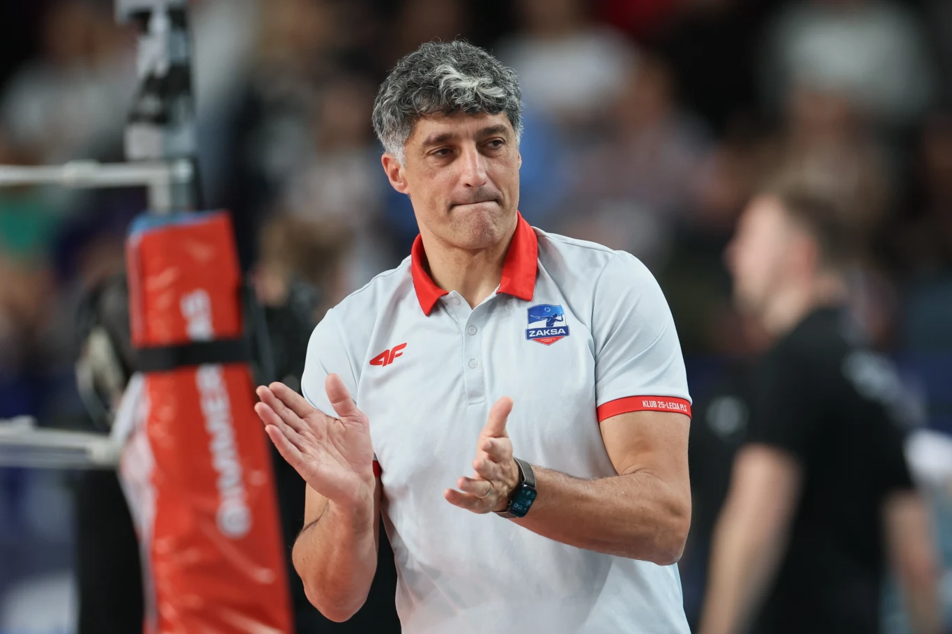 Mężczyzna w średnim wieku z krótkimi siwymi włosami ubrany w biało-czerwoną koszulkę z logo zespołu siatkarskiego ZAKSA, klaszczący w dłonie na tle rozmytego tłumu podczas wydarzenia sportowego.