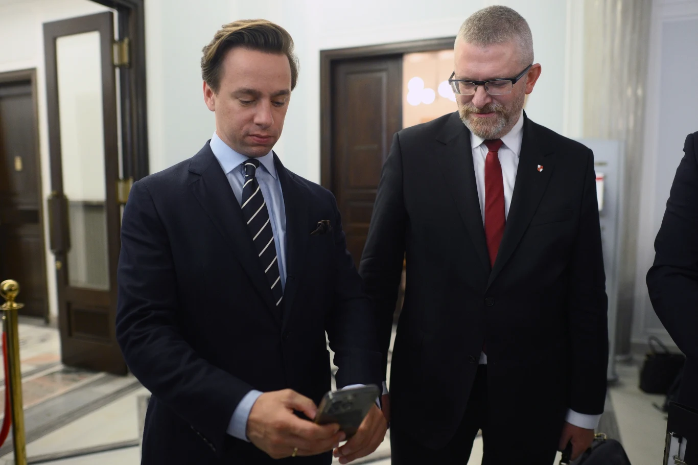 Dwóch mężczyzn w garniturach stoi w eleganckim wnętrzu, jeden z nich trzyma w ręku telefon i spogląda na ekran, drugi lekko się uśmiecha i patrzy na ekran telefonu.