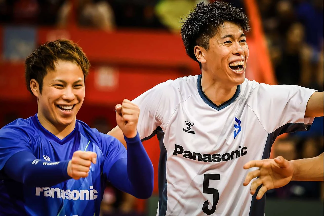 Dwóch młodych mężczyzn w sportowych strojach drużyny Panasonic świętuje sukces podczas meczu siatkarskiego, widoczne radosne emocje i uśmiechy, w tle rozmyte trybuny z kibicami.