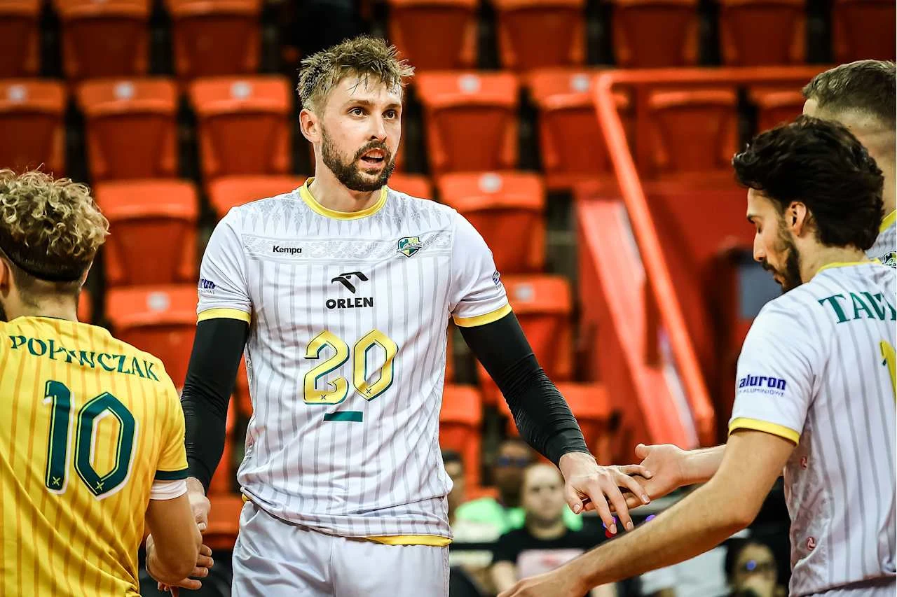 Grupa siatkarzy na boisku podczas meczu wymienia uścisk dłoni. W tle widoczne puste czerwone trybuny. Najbardziej wyeksponowany gracz nosi koszulkę z numerem 20.