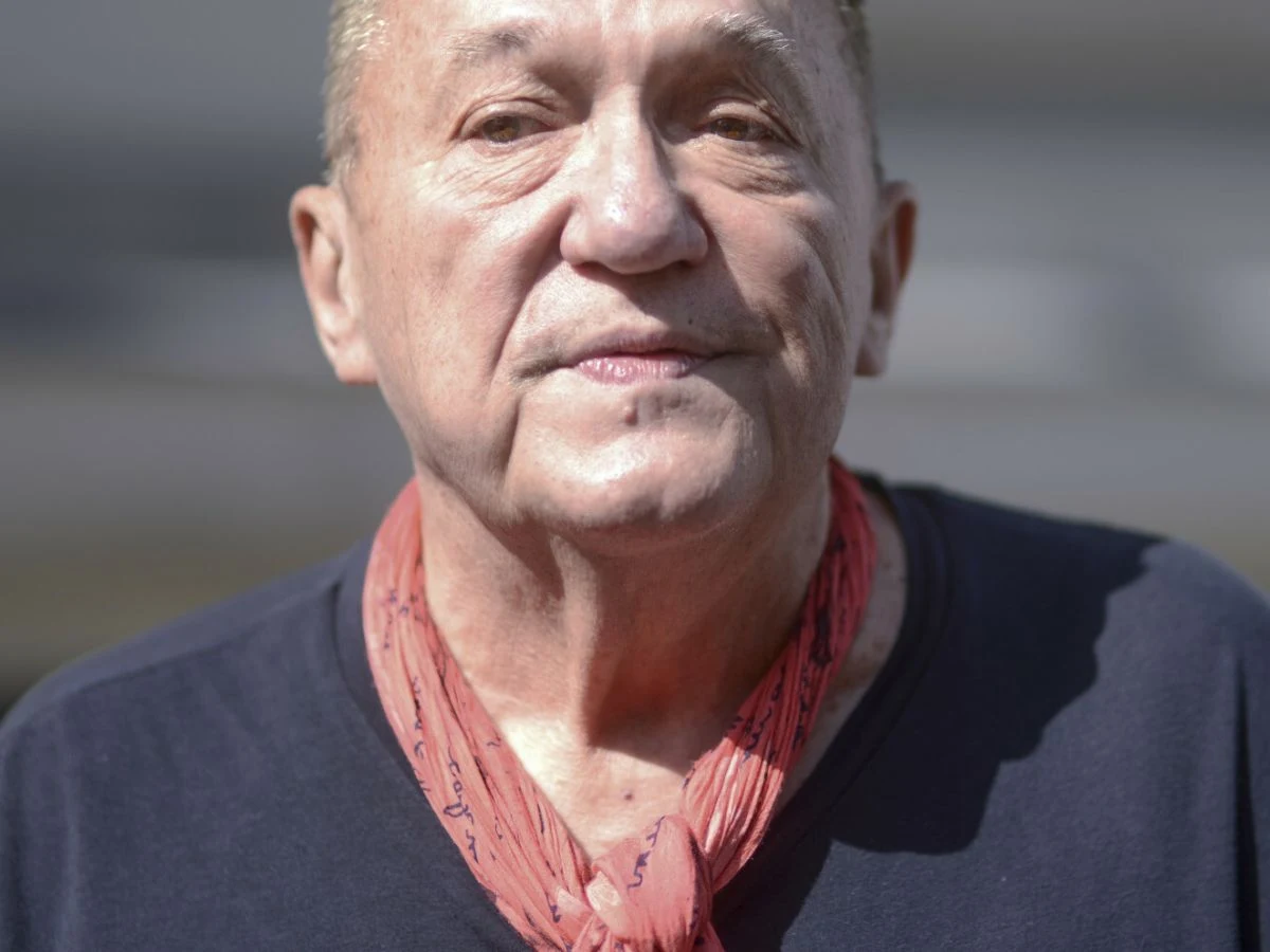 Michał Urbaniak Starszy mężczyzna w granatowej koszulce i czerwonym szaliku na szyi patrzy w obiektyw, oświetlony naturalnym światłem, na neutralnym, rozmytym tle.