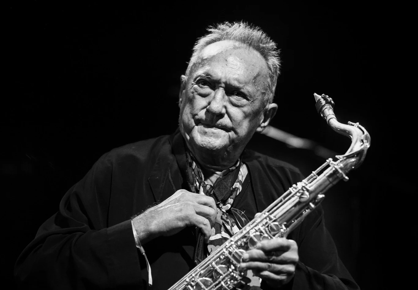 Starszy mężczyzna o skupionym wyrazie twarzy trzyma saksofon, ubrany elegancko w ciemną marynarkę i apaszkę, oświetlony punktowym światłem na ciemnym tle.