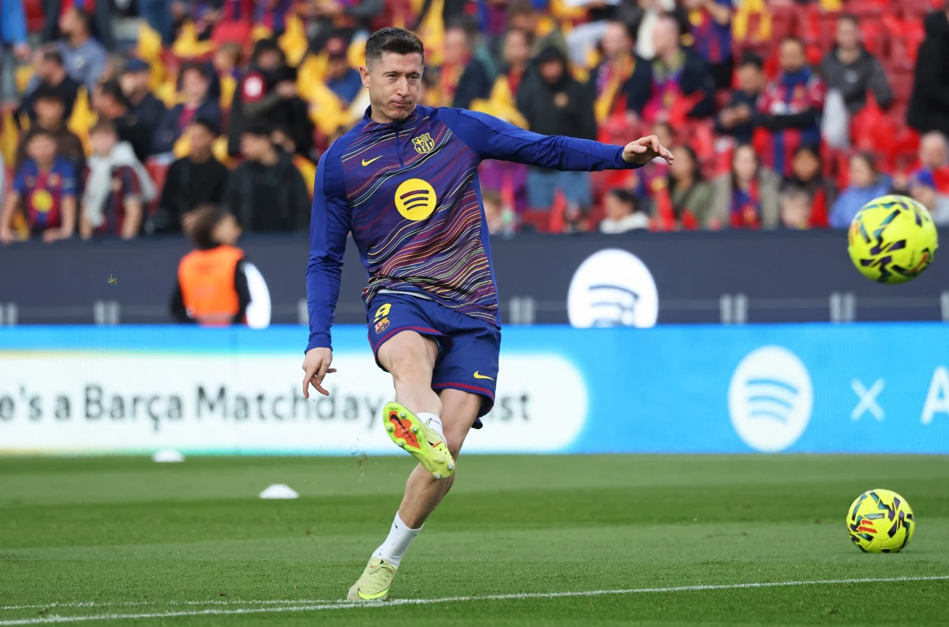 Piłkarz FC Barcelony w sportowym stroju podczas treningu na boisku, wykonujący dynamiczny kop piłki, w tle zapełnione trybuny z kibicami.