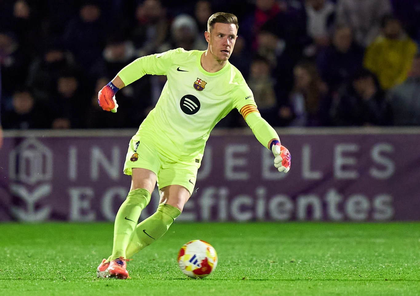 Bramkarz w jasnożółtym stroju drużyny FC Barcelona kopie piłkę podczas meczu nocą, w tle rozmazane trybuny i widoczne oświetlenie stadionu.