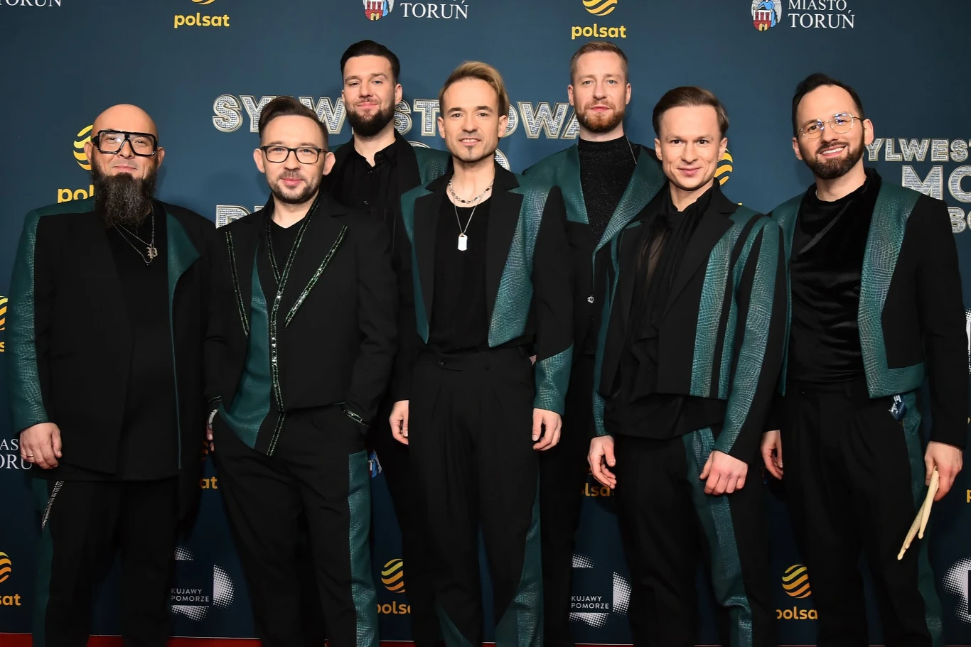 Zespół Enej po raz kolejny wystąpi na Sylwestrowej Mocy Przebojów Siedmiu mężczyzn ubranych w koordynowane, eleganckie stylizacje w odcieniach czerni i zieleni pozuje wspólnie na tle ściany z logo wydarzenia i sponsorów.