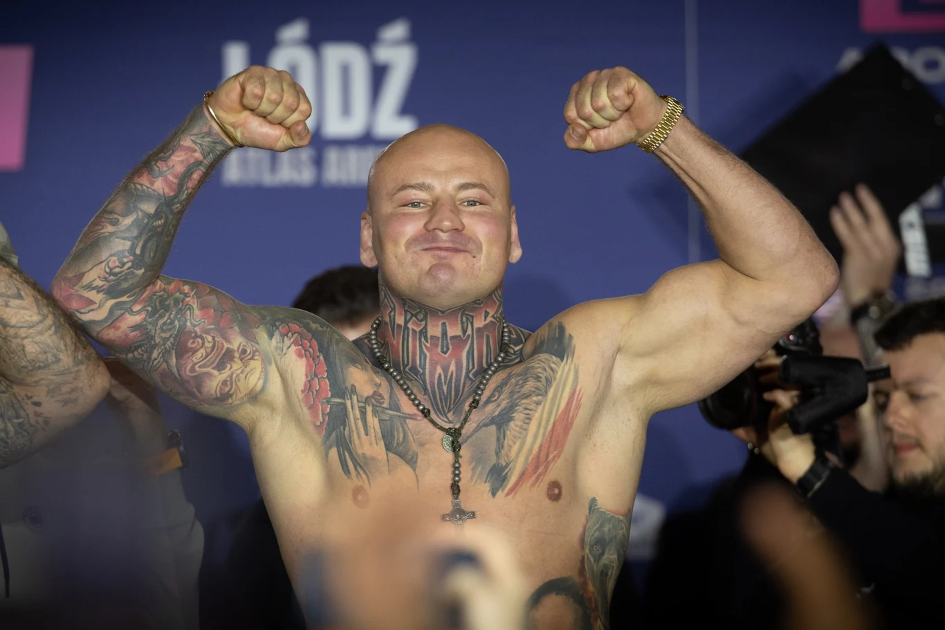 Mężczyzna z widocznymi tatuażami na klatce piersiowej oraz obu ramionach pozuje z uniesionymi, zgiętymi rękami, eksponując mięśnie i triumfalny wyraz twarzy. W tle znajduje się kilku fotografów oraz logo miasta Łódź.