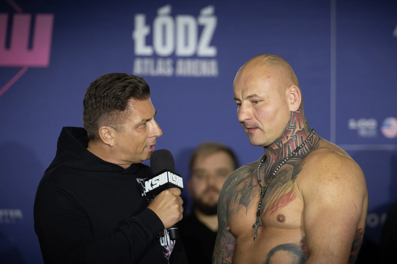 Wywiad z umięśnionym, wytatuowanym mężczyzną bez koszulki prowadzony przez reportera trzymającego mikrofon na tle ścianki z napisem Łódź Atlas Arena.