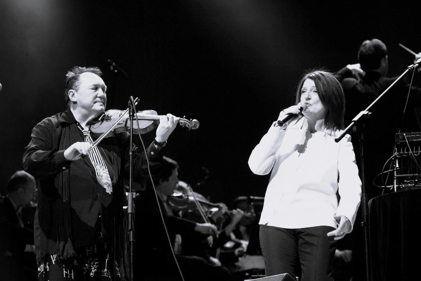 Michał Urbaniak i Urszula Dudziak wspólnie na scenie Muzyk grający na skrzypcach obok wokalistki śpiewającej do mikrofonu, oboje oświetleni reflektorami sceny, w tle widoczna orkiestra i ciemne tło sceniczne.