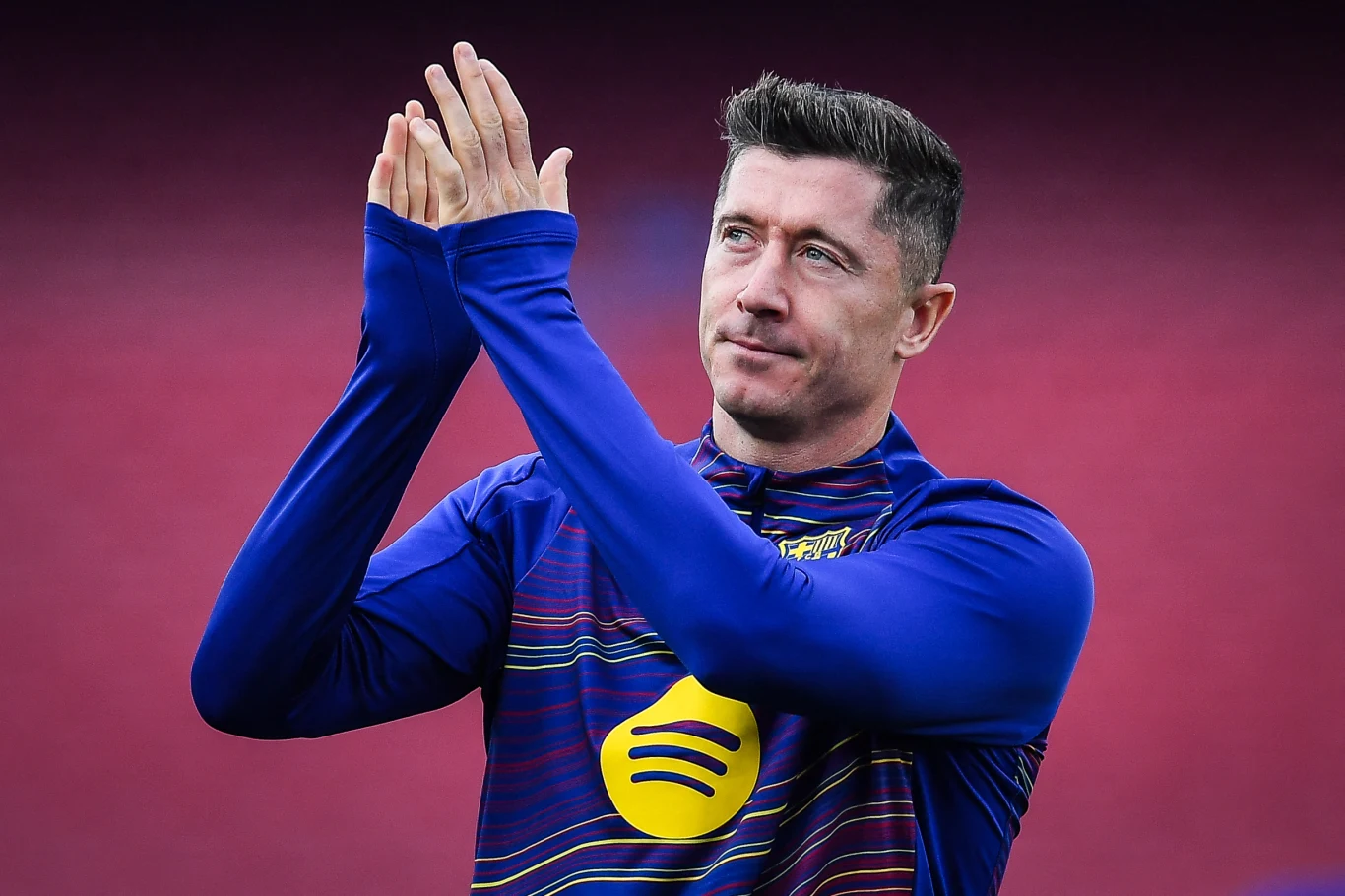 Robert Lewandowski Mężczyzna w sportowym stroju piłkarskim z logiem sponsora na piersi składa dłonie jakby dziękując lub pozdrawiając, tło w kolorze bordowym.