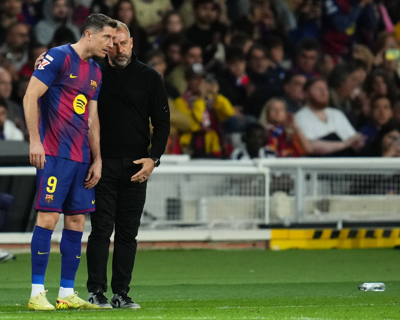 Robert Lewandowski i Hansi Flick Piłkarz w niebiesko-bordowej koszulce i trener prowadzą rozmowę przy linii bocznej boiska podczas meczu piłkarskiego, w tle widoczna publiczność na trybunach.