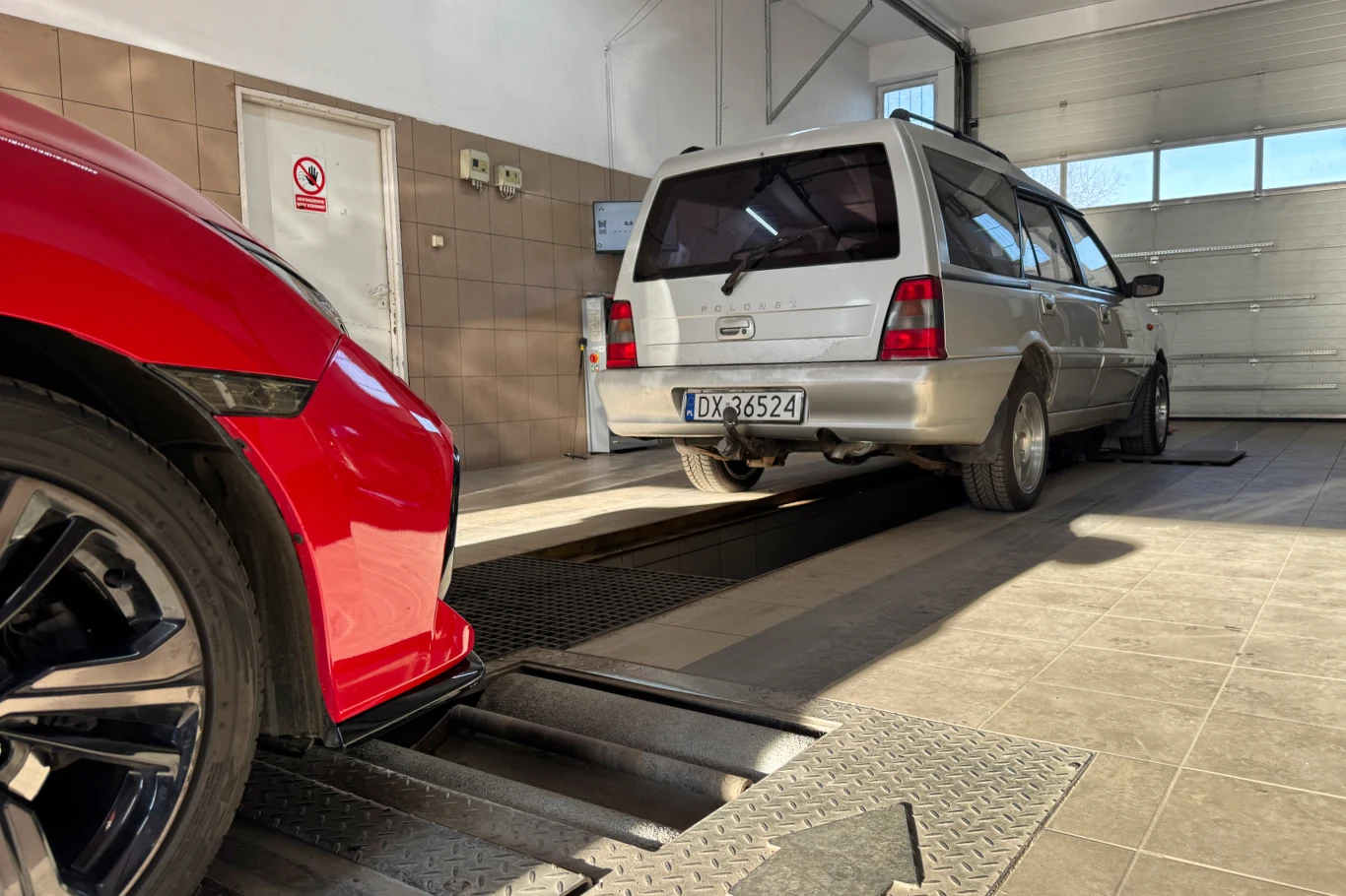 Przegląd techniczny nie zawsze kończy się pieczątką w dokumencie. Czasem kończy się przykrą decyzją w systemie Dwa samochody w pomieszczeniu warsztatowym, czerwony pojazd z przodu, srebrny kombi ustawiony przy stanowisku diagnostycznym, jasne ściany, płytki na podłodze, zamknięta brama w tle.