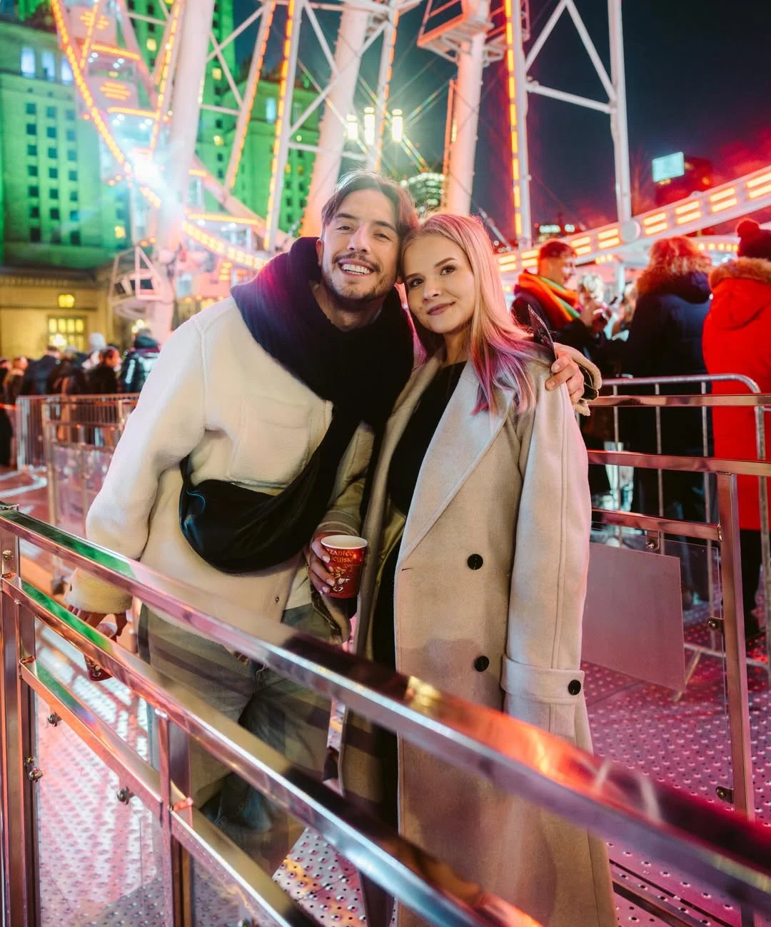 Para młodych ludzi ubrana w jasne, zimowe płaszcze stoi blisko siebie na tle oświetlonego wesołego miasteczka, uśmiechając się do aparatu, w tle widoczny tłum ludzi, iluminacje oraz fragment diabelskiego młyna.