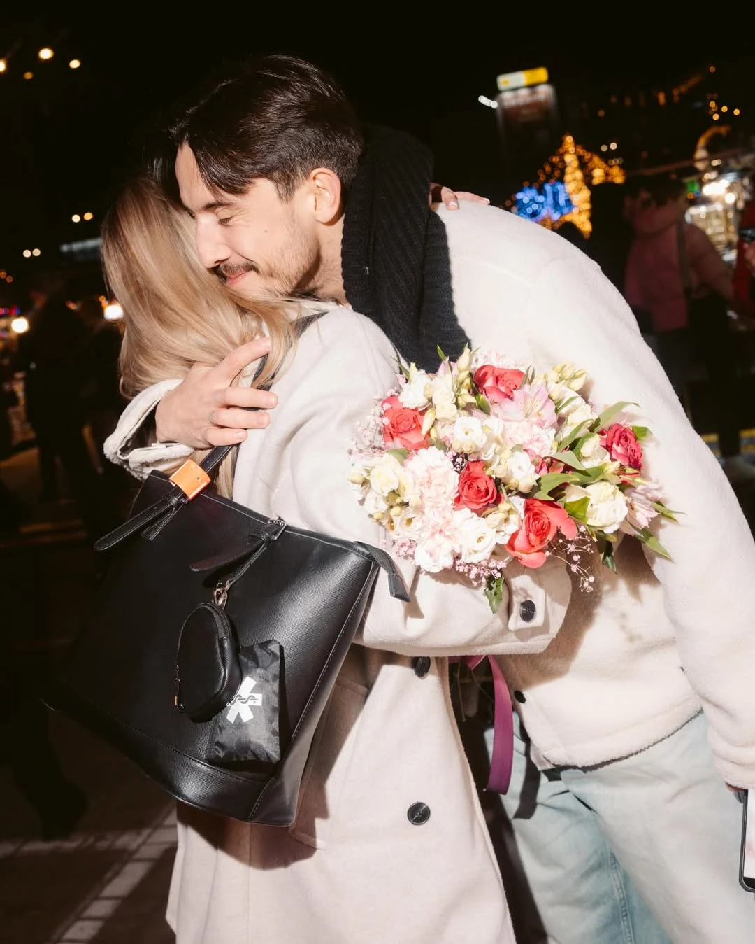 Para obejmująca się wieczorem na ulicy, mężczyzna trzyma bukiet świeżych różowych kwiatów, kobieta z czarną torebką, w tle rozświetlone dekoracje i przechodnie.