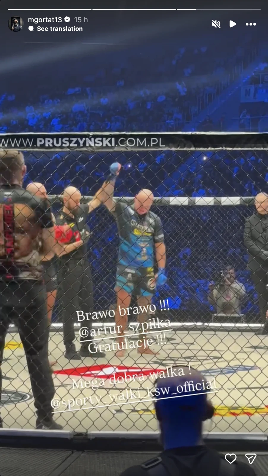 Zawodnik MMA w niebiesko-czarnych spodenkach z podniesioną ręką w geście zwycięstwa w oktagonie, obok sędzia, w tle oświetlona arena i publiczność, inni uczestnicy oraz ochrona przy siatce.