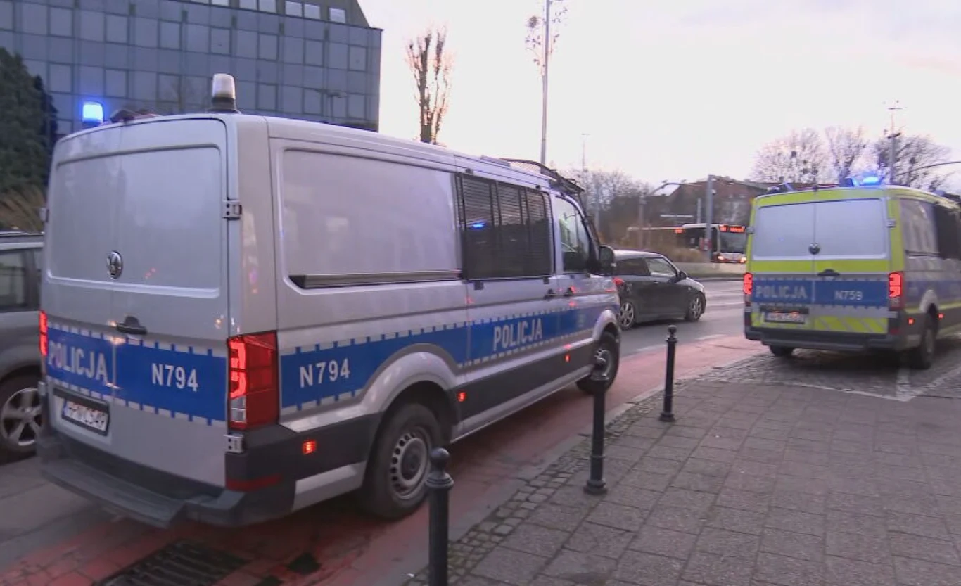 Dwa policyjne radiowozy zaparkowane na ulicy w mieście, w tle budynek biurowy oraz przechodnie, wczesny wieczór lub poranek.