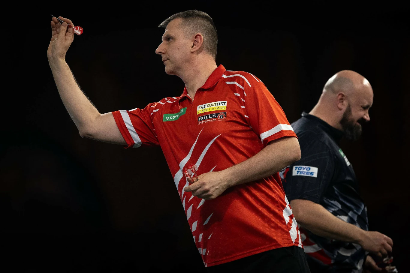 Mężczyzna w czerwonej koszulce sportowej przygotowujący się do rzutu lotką, w tle widoczny drugi zawodnik w ciemnej koszulce, scena rozgrywa się najprawdopodobniej podczas zawodów darta.