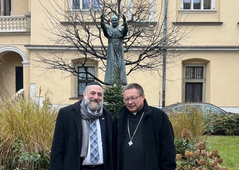 Dwóch mężczyzn stoi na tle ogrodu z rzeźbą postaci w tle oraz zabytkowym budynkiem. Jeden z mężczyzn ubrany jest w garnitur i szal, drugi w strój duchownego z krzyżem na łańcuszku. Wokół widoczne są zimowe rośliny i gałęzie drzew.