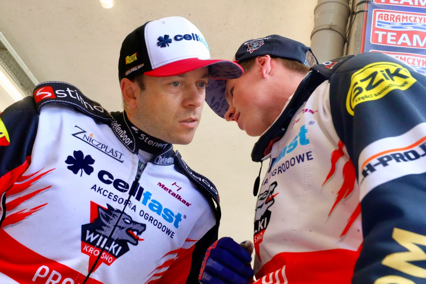 Dwóch żużlowców w kombinezonach z widocznymi nazwami sponsorów prowadzi rozmowę twarzą w twarz w otoczeniu sportowego zaplecza.
